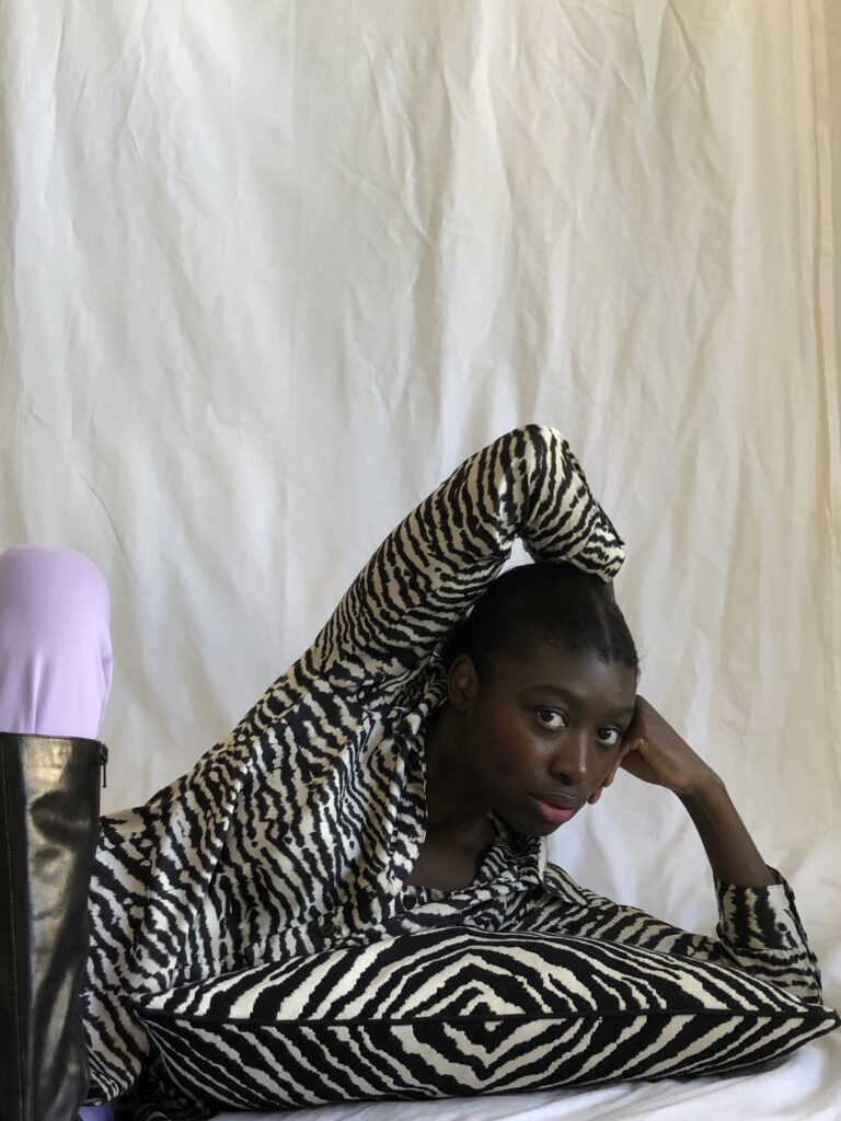 Ama Essel lies on the floor with her face resting on her hand while the other arm is raised behind her head. The background is a light-colored fabric. 